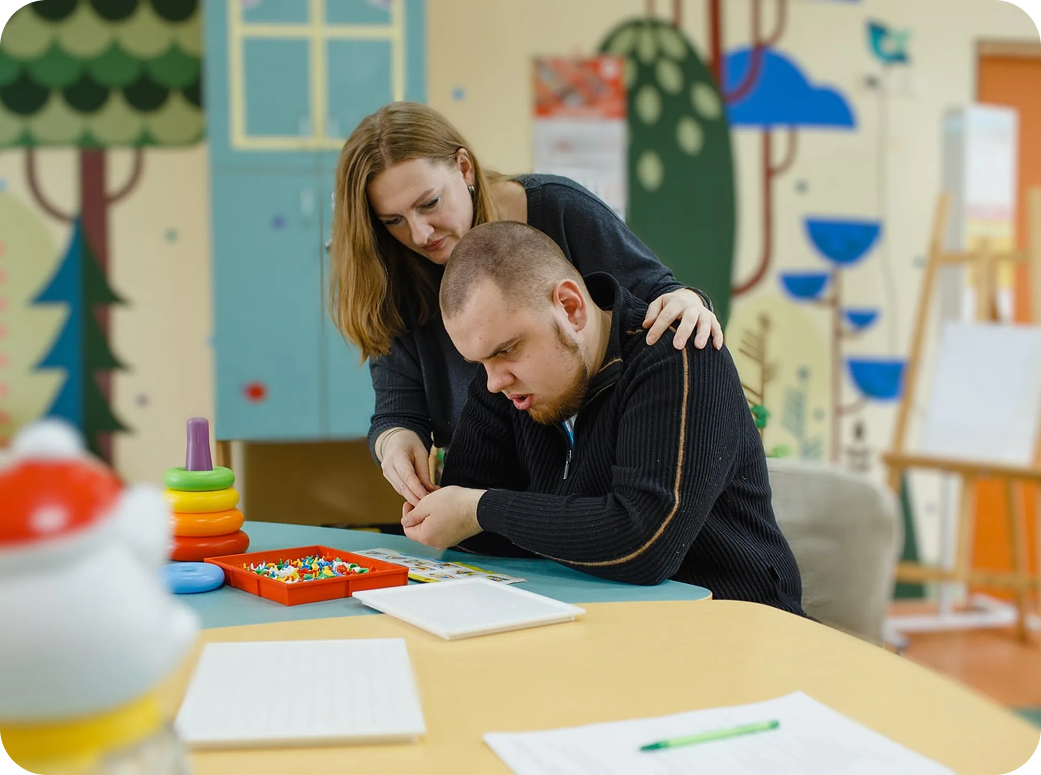 A support worker helping a young man with a writing activity at a table